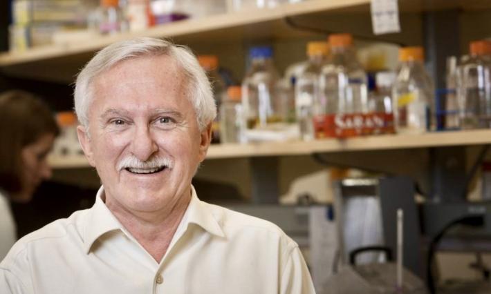 Paul Modrich, PhD is smiling while standing in his lab with bottles on shelves behind him