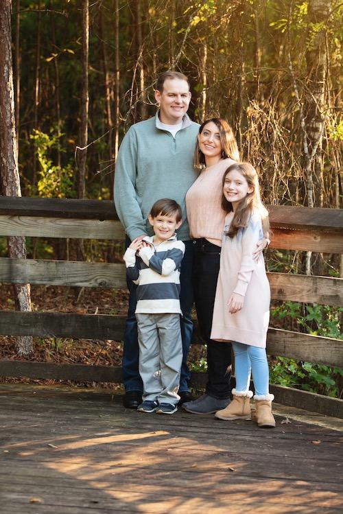 Elle Charnisky's family poses outside in front of a fence in Fall. There is her husband, Elle, and her two children, standing in front of a fence.