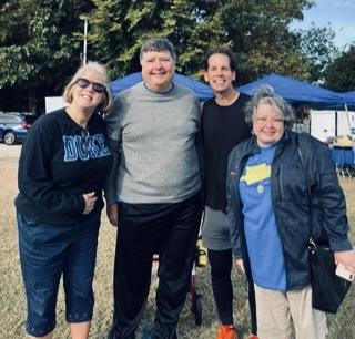 Woman in a Duke sweatshirt poses with two men and a lady with a jacket in front of blue tents