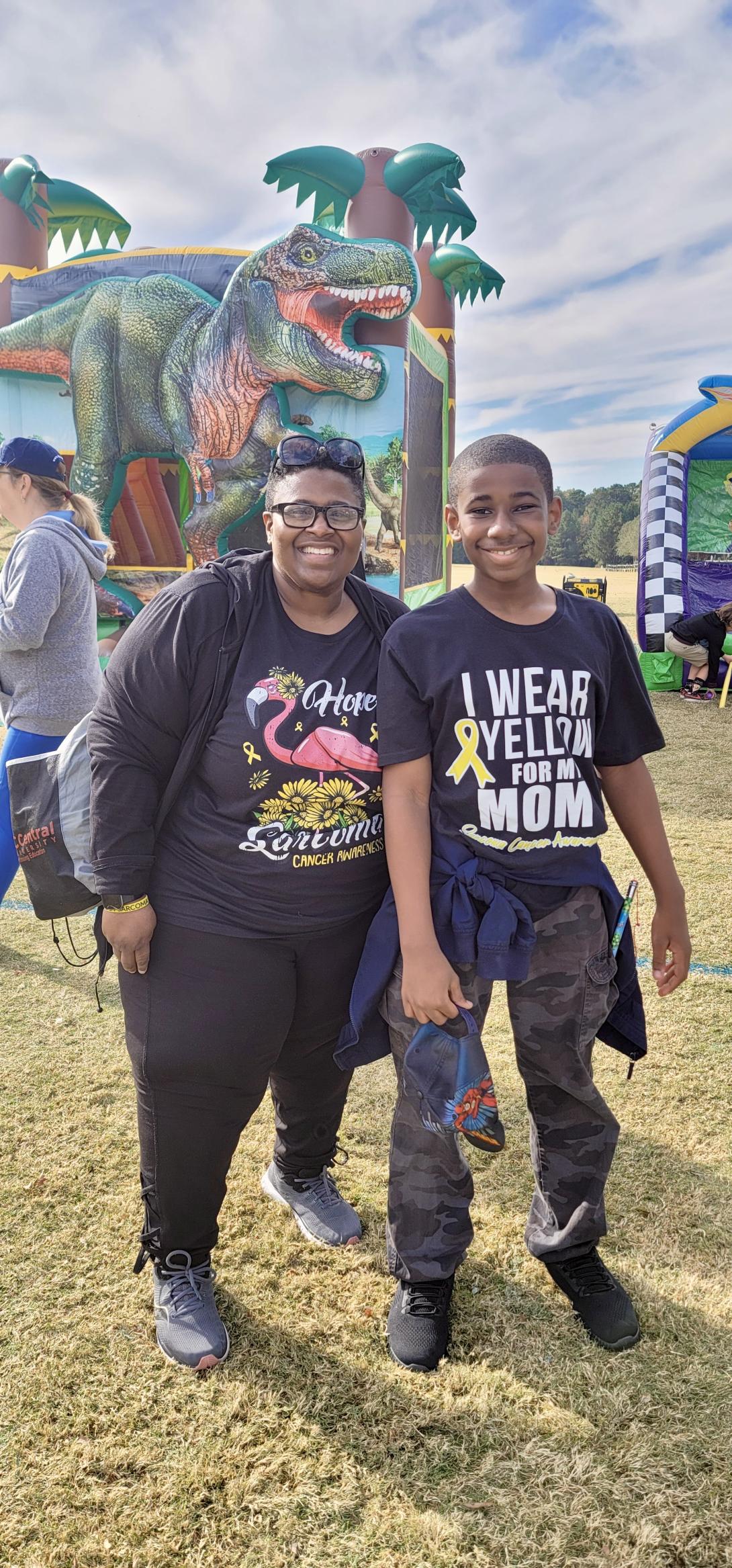 Sharon Alston with a Sarcoma Survivor shirt and her son Cason with a shirt that says "I wear yellow for my mom" stand in front of a dinosaur attraction