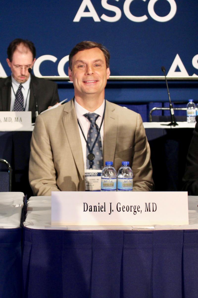 Daniel George seated at a table with his name on it and the ASCO banner in the background