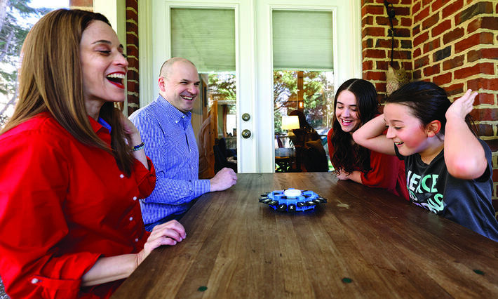 A family of four smiles and laughs at an outdoor table playing a game.