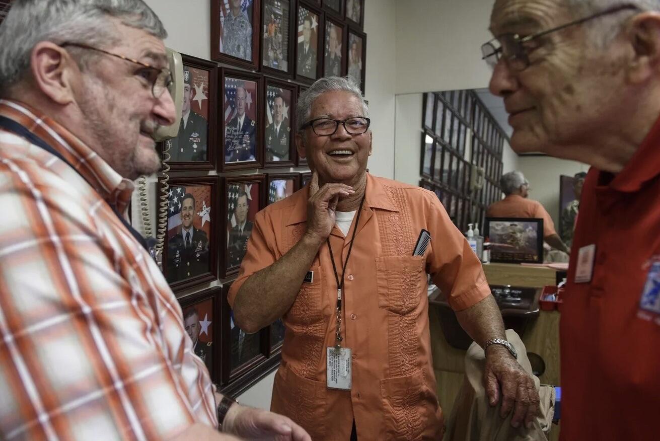 three old men laugh together in a barber shop whose wall is lined with framed photos of military generals