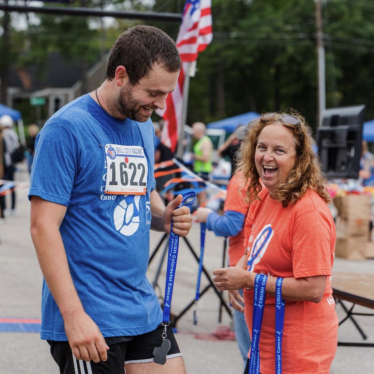 man with a blue CRUSH t-shirt and race bib receives a medal from a woman in an orange CRUSH t-shirt. Both are smiling.