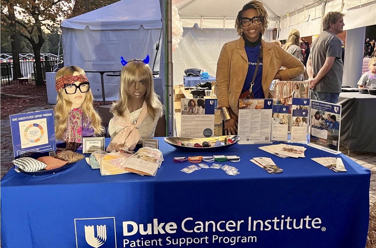 a woman in a brown suede jacket and Duke Cancer Institute shirt stands behind a table draped in a blue tablecloth with the words Duke Cancer Institute Patient Support Program. There are manniquin heads wearing wigs and glasses, papers, and small mall items on the table