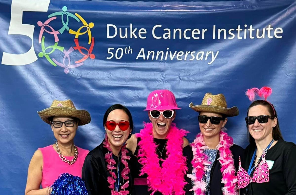 Five women dressed in pink hats, glasses, and boas, pose in front of a Duke Cancer Institute 50th Anniversary banner 