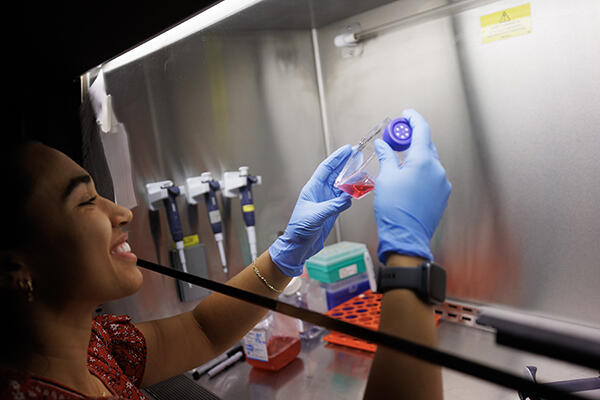 A womans wearing blue gloves holds up a vial under a fume hood.