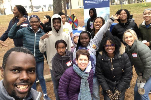 Group of about 15 people in winter coats outside on the grass smile for a selfie