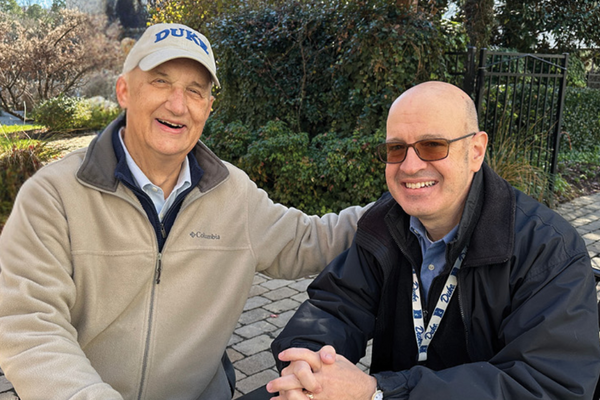 An older gentleman in a khaki colored jacket and a Duke baseball cap sits on a bench smiling with another gentleman wearing a Duke lanyard