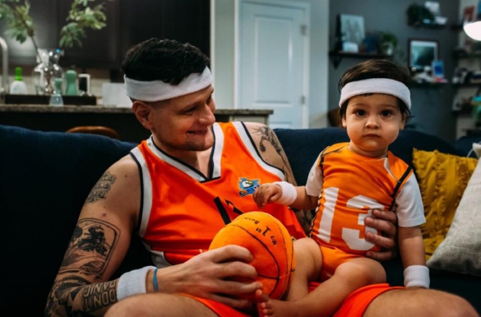 Milos Bogetic sits on the couch at home with his toddler son. They are both dressed in orange basketball jerseys and wearing white headbands. Milos is smiling at his son.