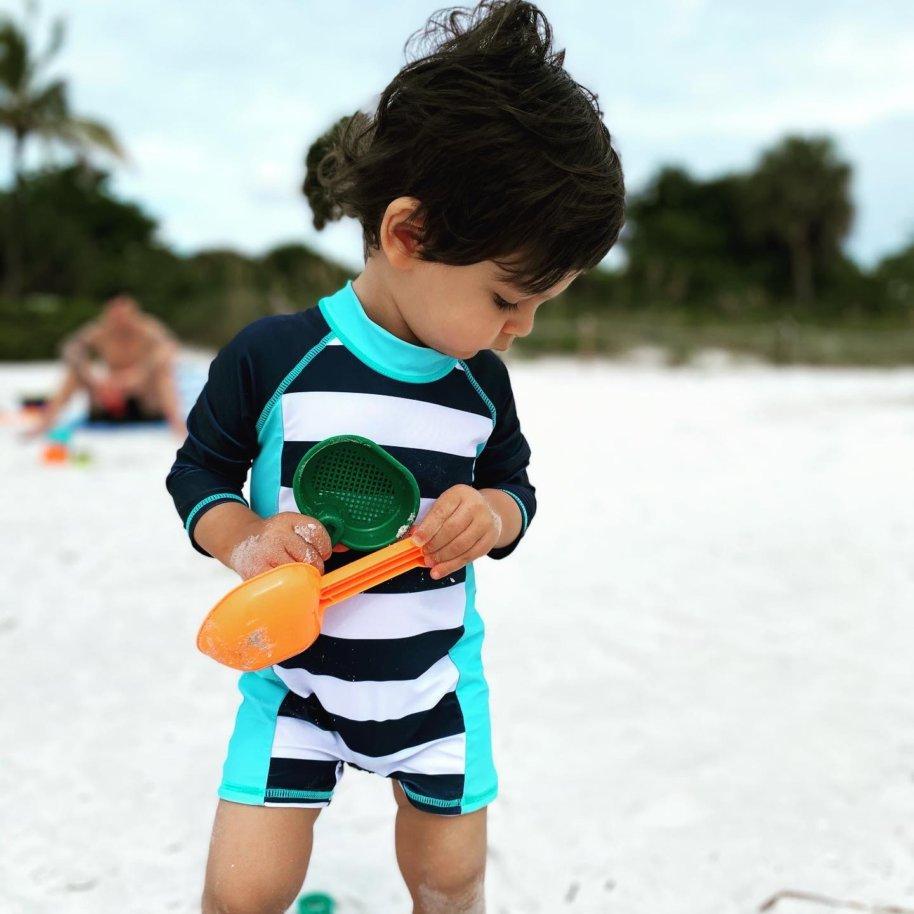 Teddy Bogetic, a toddler, plays in the sand at the beach. He is holding two shovels and looking at the ground.