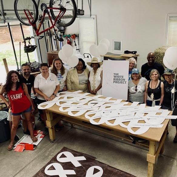 A group of 10 people stand behind a large table holding dozens of large white cardboard ribbons