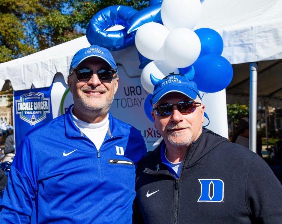 Michael Kastan and Steven Patierno pose together in sunglasses, blue Duke sportswear, and caps in front of a white tent with blue and white balloons