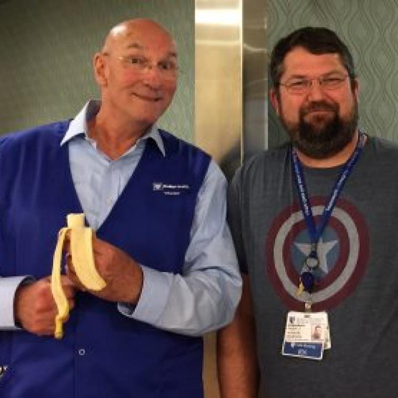 man in duke blue volunteer vest holding a banana stands with man with Duke ID and a star on his shirt