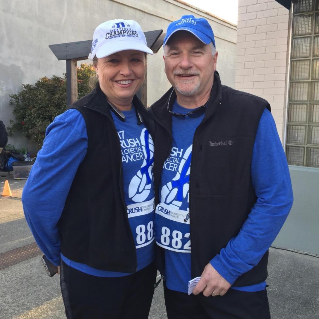 A woman and a man in long sleeve blue t-shirts, black vests, and baseball caps pose outside