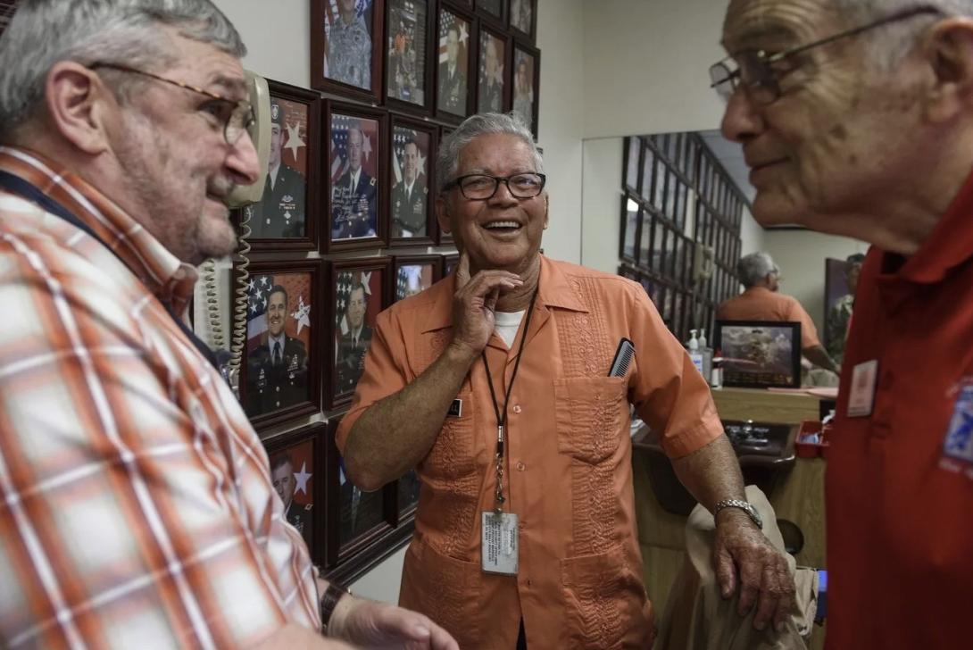 three old men laugh together in a barber shop whose wall is lined with framed photos of military generals