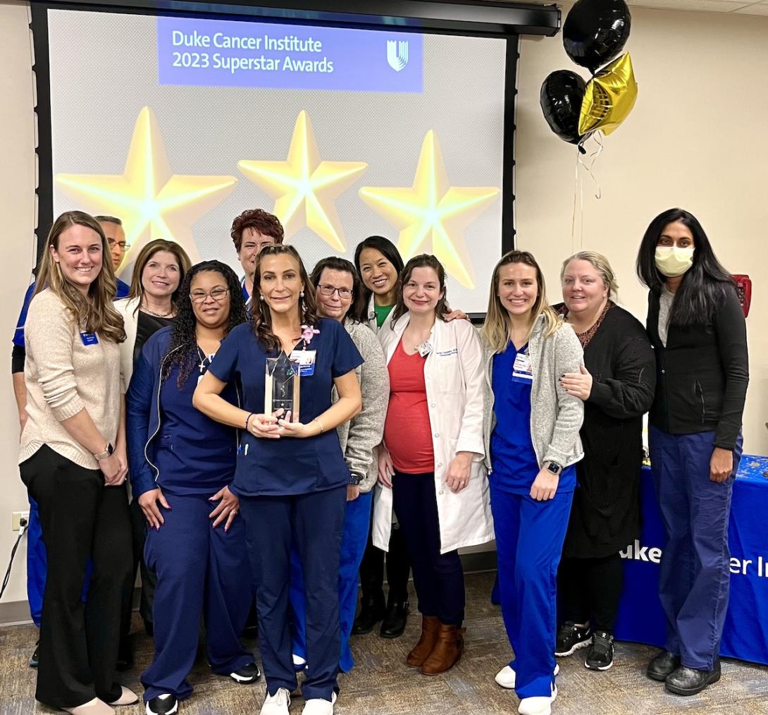 woman standing with a glass trophy and 11 others surrounding her, posing as a group. A large screen displaying three yellow stars and reading "Duke Cancer Intitute 2023 Superstar Awards" is behind them.